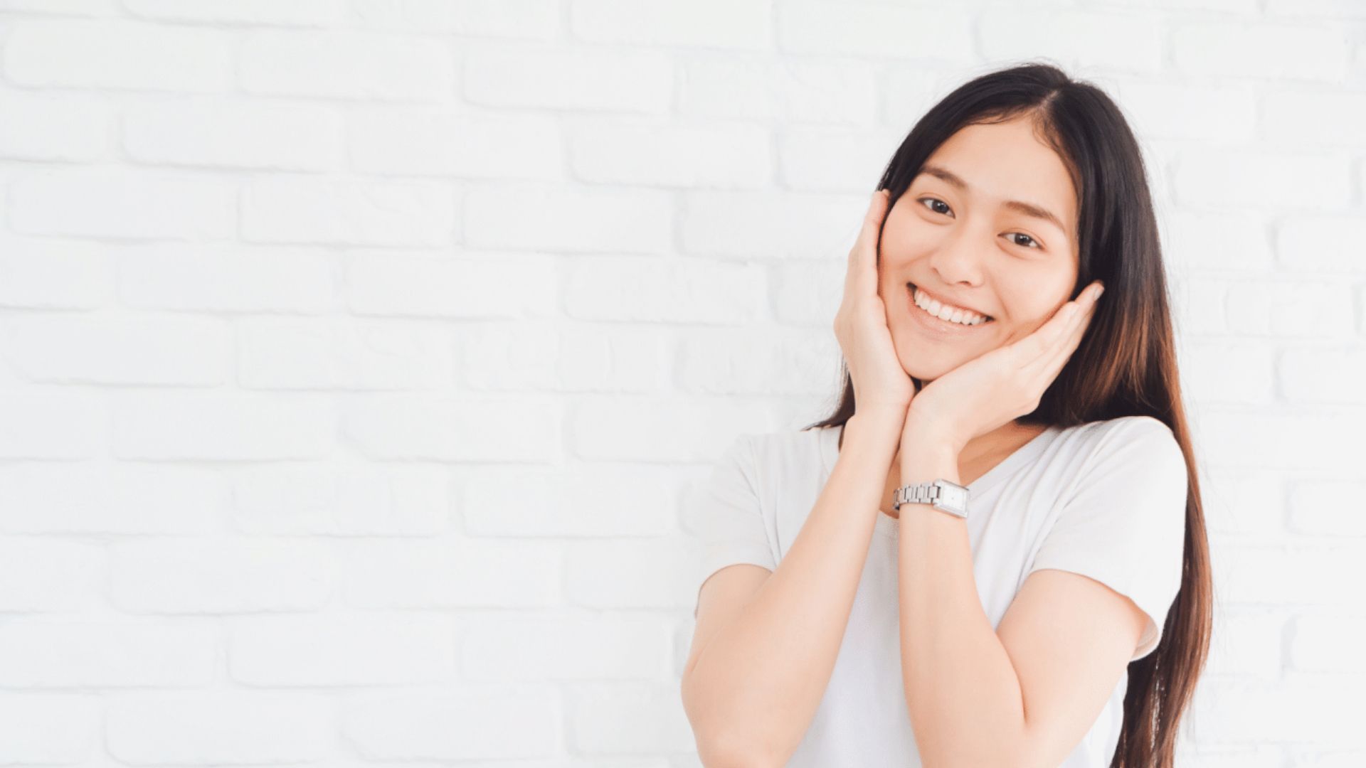 A long-haired young girl wearing a white shirt has her hands cupping her face. Background is a while brick wall.