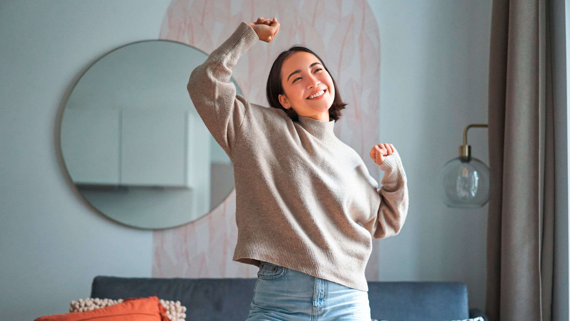 A person is dancing in a stylish living room with a gray sofa, patterned cushion, orange throw, and a round wall mirror, wearing a beige sweater and light blue jeans.