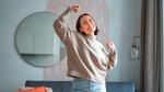 A person is dancing in a stylish living room with a gray sofa, patterned cushion, orange throw, and a round wall mirror, wearing a beige sweater and light blue jeans.