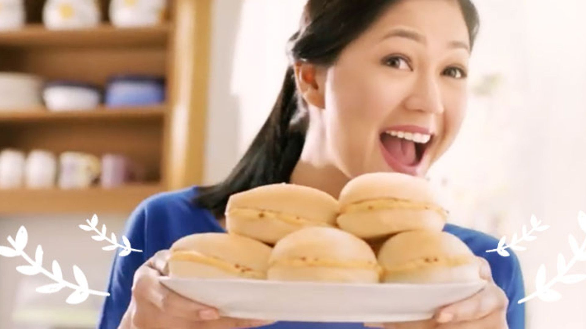 Woman smiling and holding a plate with a stack of round cream-filled buns in a bright kitchen setting.