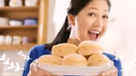 Woman smiling and holding a plate with a stack of round cream-filled buns in a bright kitchen setting.