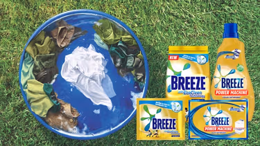 A laundry tub with green, brown and white clothes is placed on top of outdoor grass. Next to it are four Breeze products.