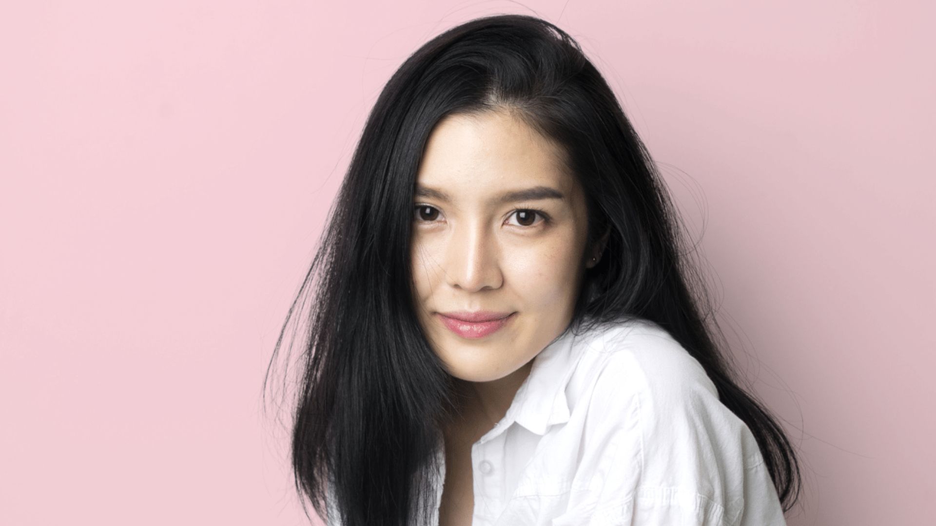 A long-haired young girl wearing a white shirt has her hands cupping her face. Background is a while brick wall.
