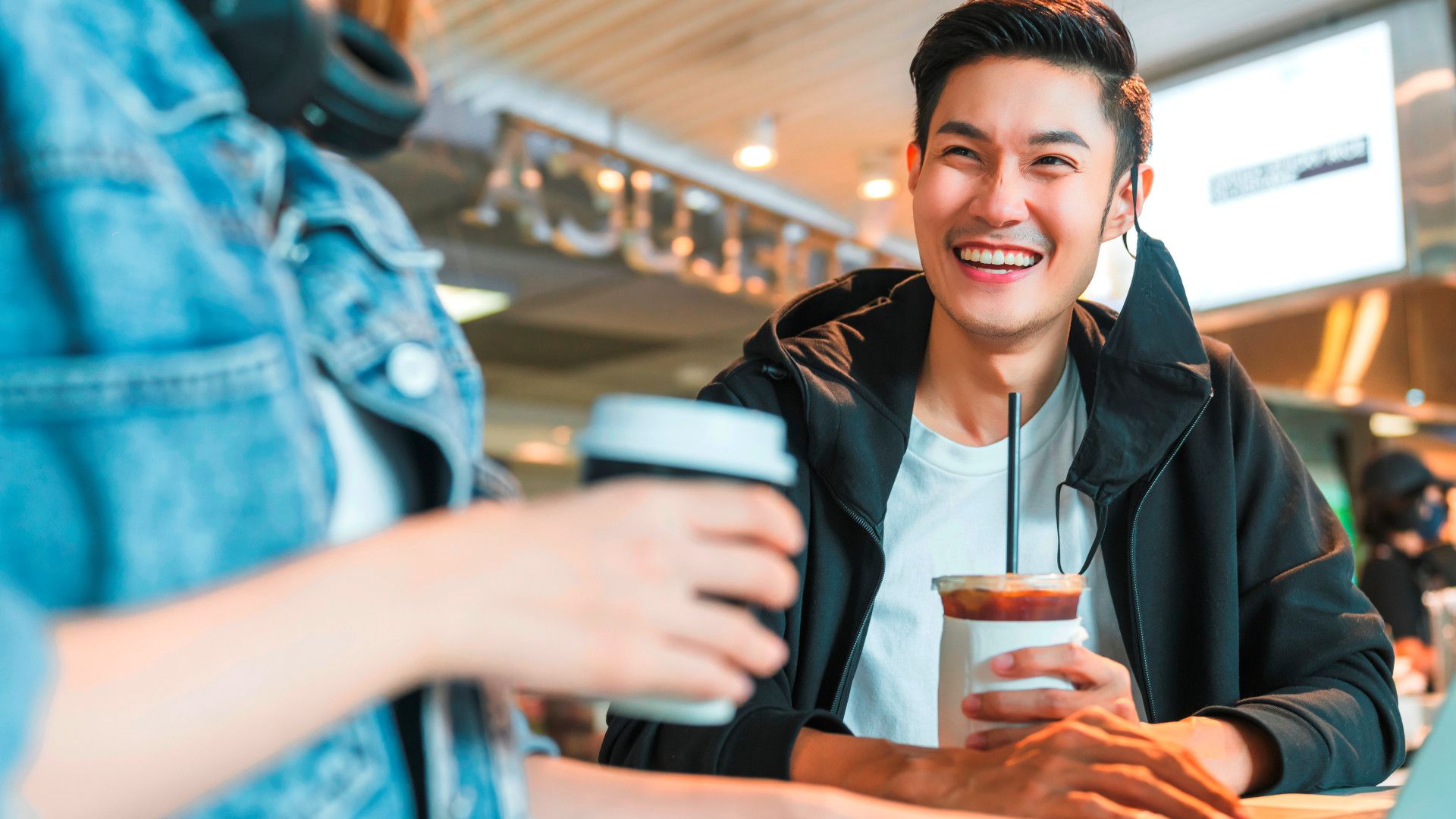 A man in a black hoodie and holding a drink, sitting at a counter, and smiling at a friend.
