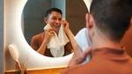 A man smiles while drying his face with a towel in front of a round, illuminated mirror.