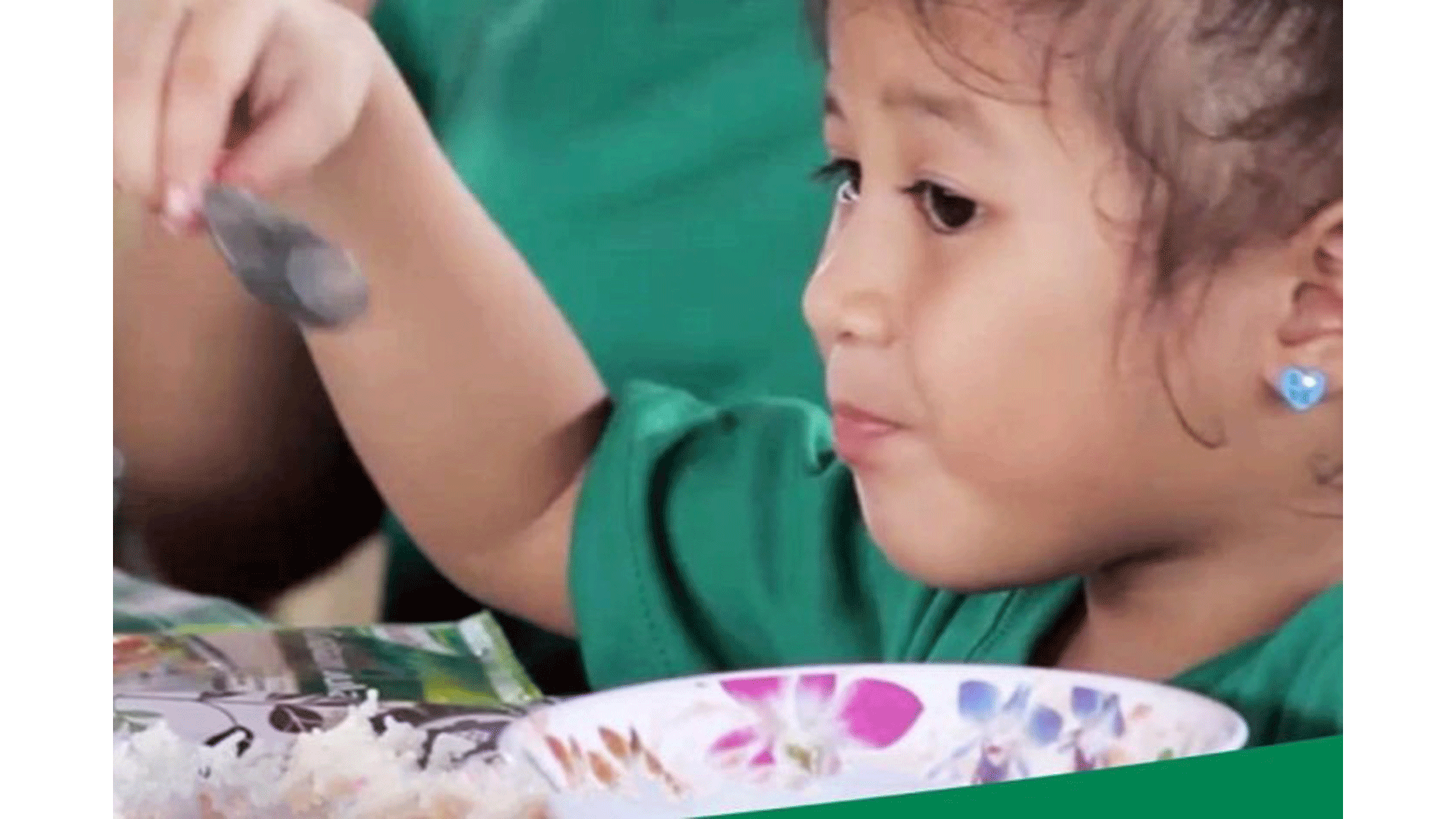 A female child eats while holding a spoon. In front of her is rice and a soup bowl
