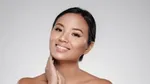 A Filipina woman is smiling at the camera while gently placing her hands near her cheek and neck.