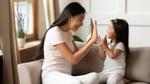 A woman and young girl sitting on a couch indoors, giving each other a high five in a bright living room.