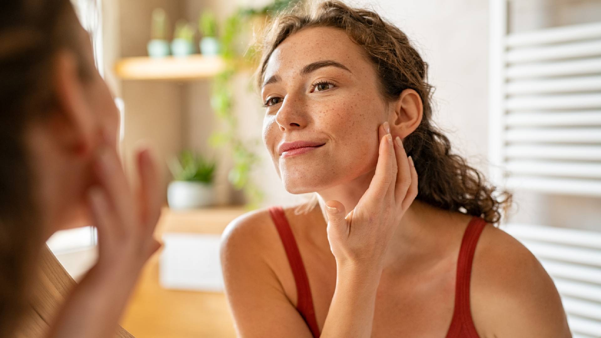 Woman in a bathroom, smiling at her reflection in the mirror. She has her hand to her face and is wearing a red top.